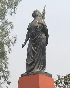 Bronze statue of Matangini Hazra on a red plinth in Kolkata, standing with one arm outstretched, trees and open sky behind.